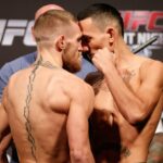 Conor McGregor falando em entrevista coletiva de boxe com um cinturão de campeão e produtos em uma mesa à sua frente.