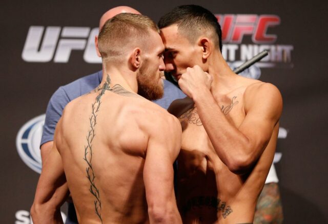 Conor McGregor falando em entrevista coletiva de boxe com um cinturão de campeão e produtos em uma mesa à sua frente.