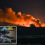 Vista noturna do horizonte de uma cidade com grandes nuvens de fumaça laranja subindo para o céu por trás dos edifícios.