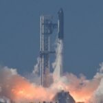 Um foguete espacial voa para cima em um céu azul claro, deixando um enorme rastro cinza atrás de si em uma faixa cada vez maior.