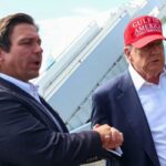 O governador da Flórida, Ron DeSanti, dá as mãos ao presidente dos EUA, Donald Trump, que usa um chapéu vermelho que diz