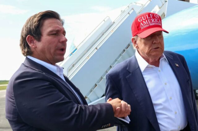 O governador da Flórida, Ron DeSanti, dá as mãos ao presidente dos EUA, Donald Trump, que usa um chapéu vermelho que diz 
