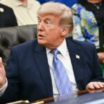 Donald Trump em terno azul marinho e gravata listrada azul clara gesticula com a mão direita enquanto fala.