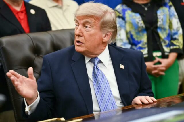Donald Trump em terno azul marinho e gravata listrada azul clara gesticula com a mão direita enquanto fala.