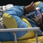 Um homem com um casaco de escalada azul bebendo uma sopa de laranja em uma tigela, sendo ajudado por um homem com um casaco preto