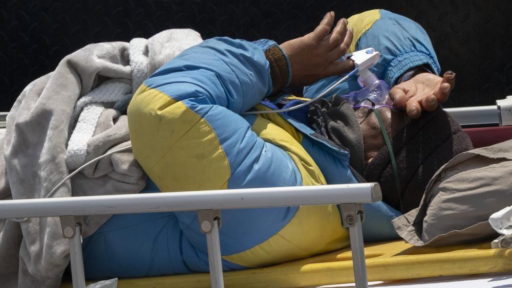 Um homem com um casaco de escalada azul bebendo uma sopa de laranja em uma tigela, sendo ajudado por um homem com um casaco preto
