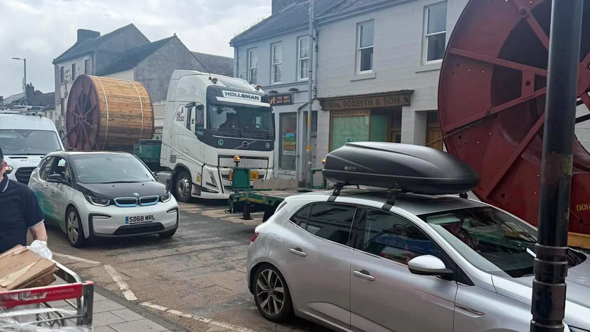 Uma foto de obras rodoviárias em Sanquhar com andaimes na estrada próximo ao histórico Museu Tolbooth à esquerda. Há uma placa de estrada fechada e semáforos e um homem de macacão laranja ao telefone e outro com jaqueta de alta visibilidade e capacete conversando com um membro do público