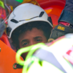 a imagem mostra metade do rosto dos meninos – o resto está obscurecido pelas equipes de resgate. Ele está usando um capacete branco e tem cabelo escuro e encaracolado.