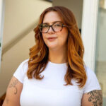 Jess King, uma mulher com longos cabelos ruivos e óculos, está parada em frente a um balcão de cozinha com eletrodomésticos e um bloco de facas. Ela está vestindo uma blusa branca de mangas curtas e calças marrons e tem tatuagens pretas visíveis na parte superior dos braços.