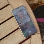 Mão segurando um smartphone com elementos interativos destacados na tela contra um fundo gradiente rosa com ícones brilhantes.