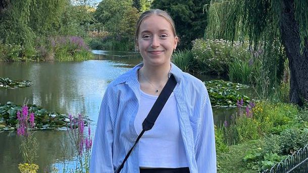 Libby sorrindo para a câmera em frente a um lago com nenúfares, plantas e flores. Ela tem cabelo loiro amarrado para trás e veste uma camisa listrada azul e branca com uma regata branca por baixo. Ela também está usando uma bolsa preta e um colar de prata simples com um pequeno pingente.