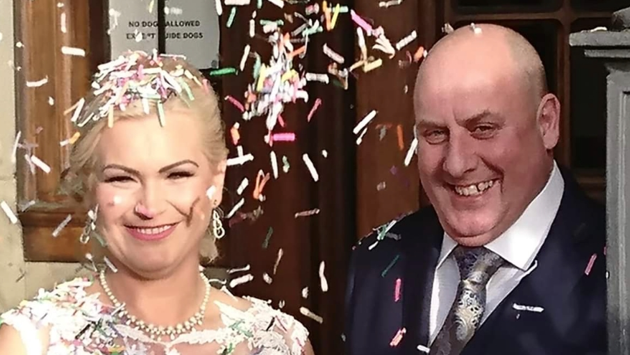 Anthony Webb e sua esposa Ewelina no dia do casamento. Os dois estão sorrindo e olhando para a câmera, com confetes em volta de seus rostos. Ele é careca e usa um terno azul, seu cabelo loiro está preso para trás e ela usa um colar de cascas brancas e um vestido de noiva.