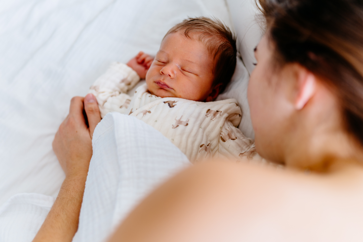Close-up of newborn baby peacefully sleeping next to mother, wrapped in a soft white blanket.