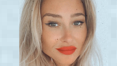 Joanna Derkacz sorri para a câmera em uma foto em close. Ela tem longos cabelos loiros e usa maquiagem nos olhos e batom vermelho.