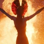 An alien warrior holds two blades up against a background of fire