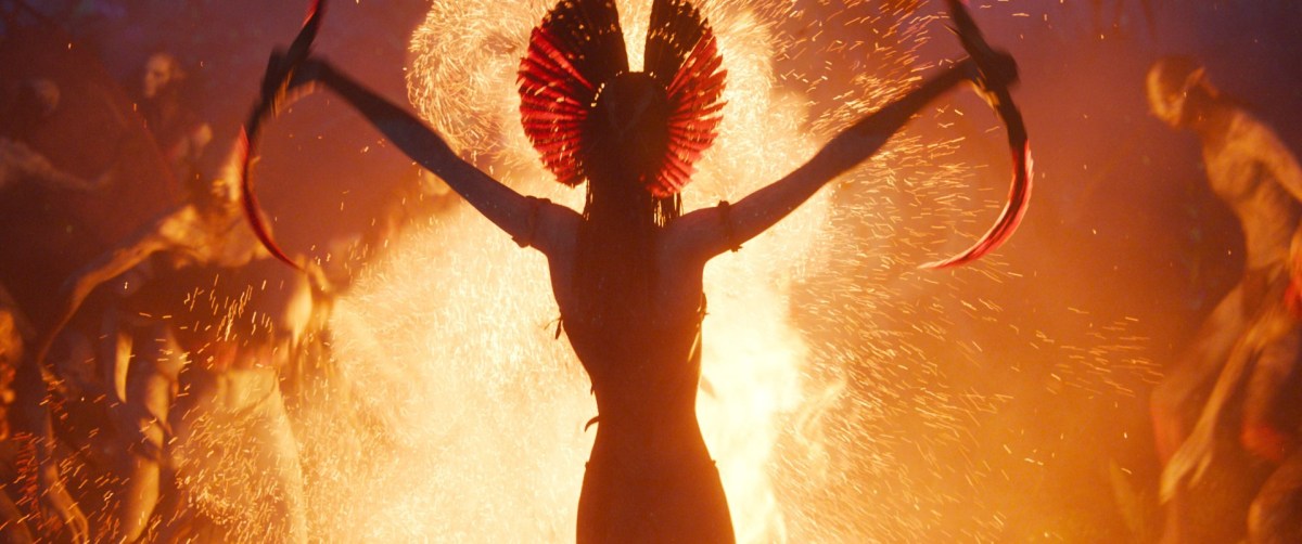 An alien warrior holds two blades up against a background of fire