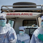 Dois profissionais de saúde são fotografados usando equipamento de proteção azul