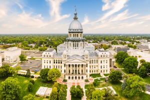 Vista aérea do edifício do Capitólio do Estado de Illinois em Springfield, Illinois, mostrando o marco histórico do governo, a cúpula icônica, os jardins paisagísticos e o horizonte do centro da cidade sob um céu azul brilhante. Projeto de lei orçamentária de Illinois visa mercados de previsão esportiva com imposto exclusivo sobre transações