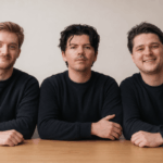 Three caucasian men in black long-sleeved crewneck shirts sit at a desk against a beige background.