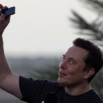 Elon Musk vestindo uma camiseta preta e segurando o telefone acima da cabeça.