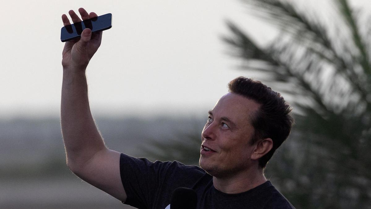 Elon Musk vestindo uma camiseta preta e segurando o telefone acima da cabeça.