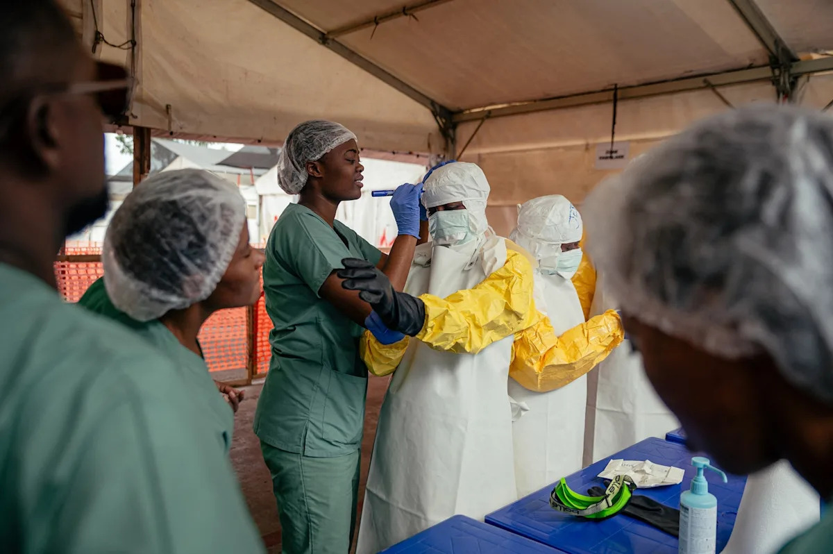 Jospin Mwisha/AFP via Getty Images - FOTO: Profissionais de saúde colocam equipamentos de proteção individual na área de curativos sob a supervisão de especialistas antes de examinarem os pacientes na enfermaria de isolamento do Centro de Tratamento de Ebola em Munigi, em 2 de junho de 2026.