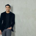 A well-dressed man leans against a neutral colored wall