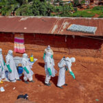 Gradel Muyisa Mumbere/Reuters - FOTO: Trabalhadores da Cruz Vermelha usando equipamentos de proteção individual caminham em formação enquanto desinfetam o chão do lado de fora da casa de um homem não identificado que morreu de Ebola em Mongbwalu, República Democrática do Congo, em 24 de maio de 2026.