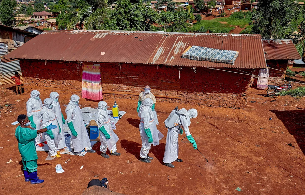 Gradel Muyisa Mumbere/Reuters - FOTO: Trabalhadores da Cruz Vermelha usando equipamentos de proteção individual caminham em formação enquanto desinfetam o chão do lado de fora da casa de um homem não identificado que morreu de Ebola em Mongbwalu, República Democrática do Congo, em 24 de maio de 2026.