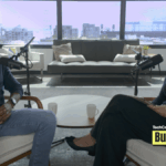 TechCrunch Build Mode podcast host Isabelle Johannessen sits across from venture capitalist Charles Hudson in a bright podcast studio. Both are smiling toward the camera, seated in armchairs with microphones positioned between them. A coffee table with two mugs sits in the foreground, while large windows behind them overlook the San Francisco skyline. The TechCrunch Build Mode logo appears in the bottom-right corner.