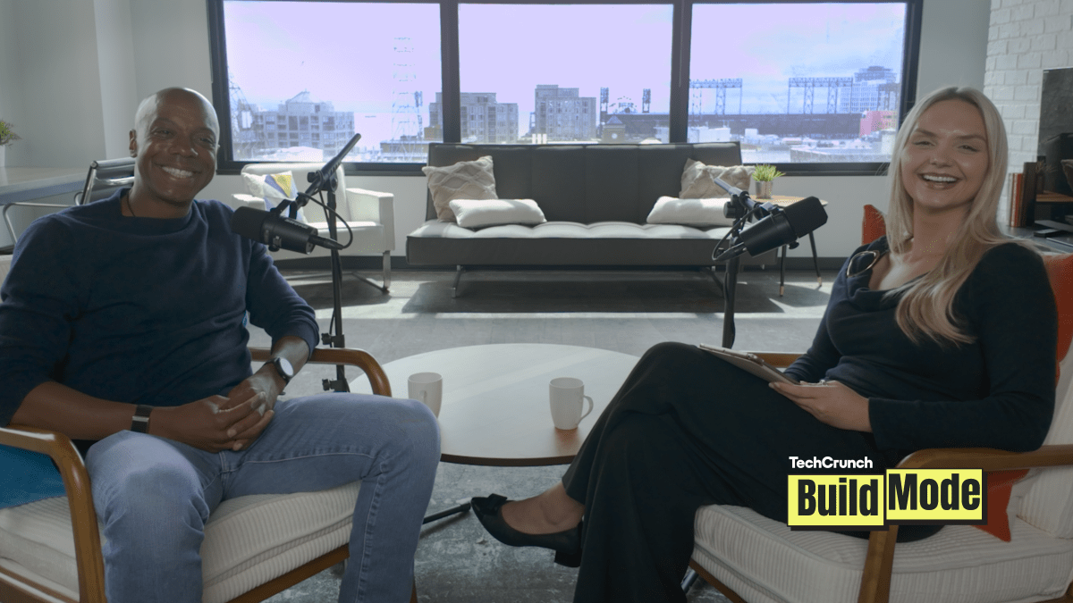 TechCrunch Build Mode podcast host Isabelle Johannessen sits across from venture capitalist Charles Hudson in a bright podcast studio. Both are smiling toward the camera, seated in armchairs with microphones positioned between them. A coffee table with two mugs sits in the foreground, while large windows behind them overlook the San Francisco skyline. The TechCrunch Build Mode logo appears in the bottom-right corner.