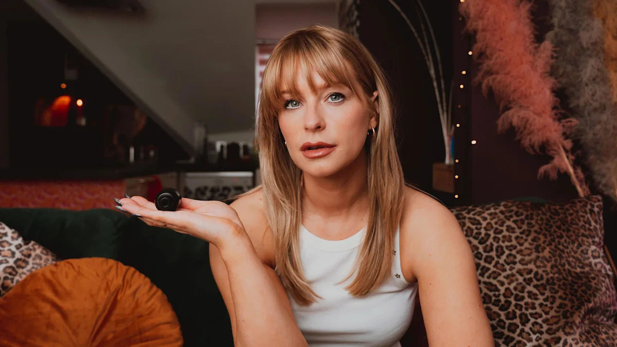 Jess sentada em seu apartamento olhando para o telefone. Uma TV em um armário pode ser vista atrás dela, com uma parede turquesa com obras de arte emolduradas à sua esquerda.
