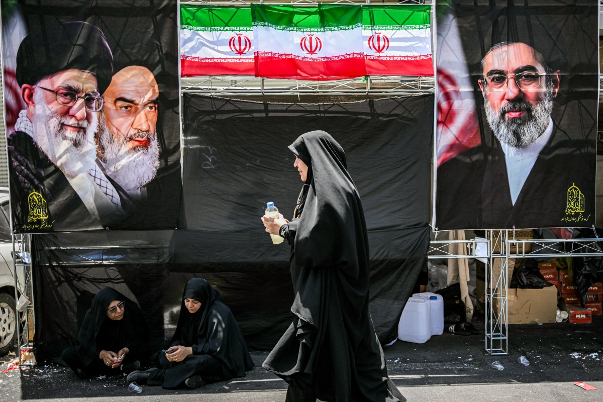 A woman walks past images of Iran's current supreme leader Mojtaba Khamenei (R) and his predecessors, Ali Khamenei (L) and the late Ruhollah Khomeini (2nd-L) on the way to the Grand Mosalla in Tehran on July 5, 2026, for the funeral ceremonies of Ali Khamenei. Photo by Ozan KOSE / AFP via Getty Images.