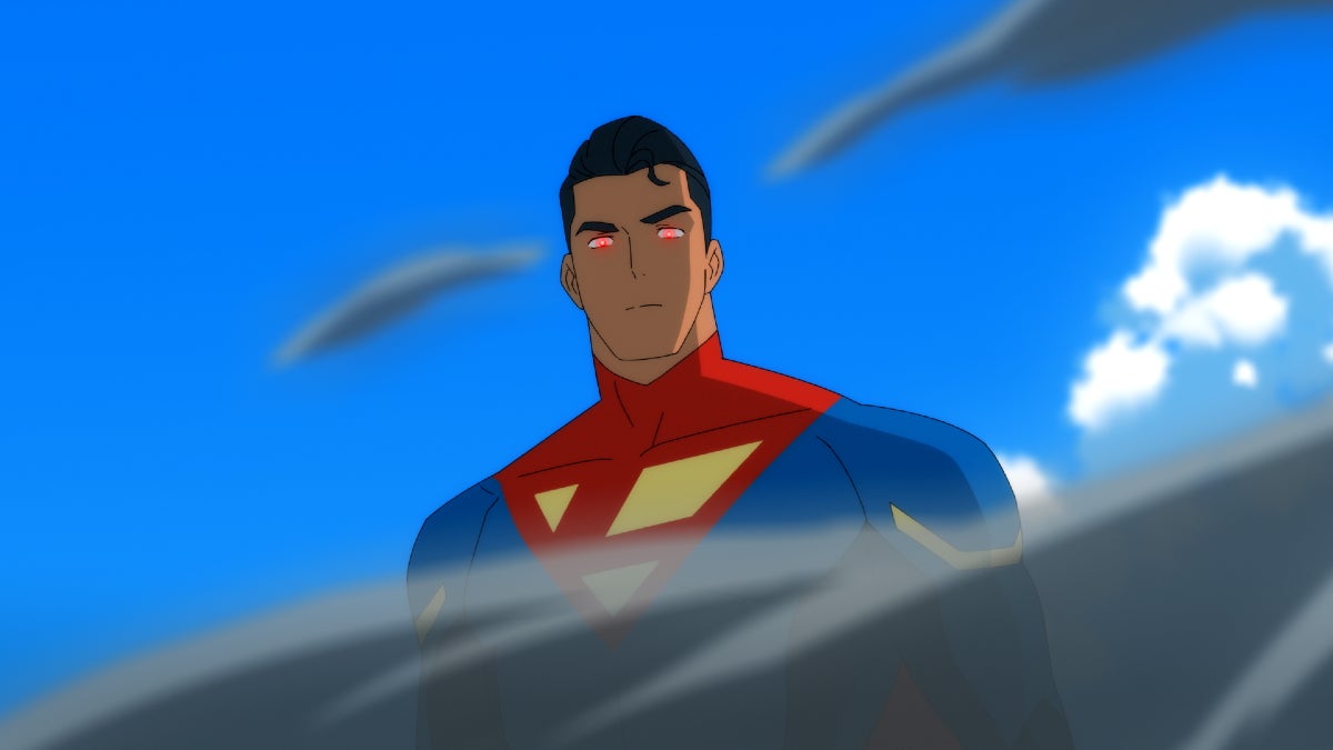 Um jovem latino veste uma fantasia de super-herói azul neon, o azul iluminado, com o capacete abaixado, mostrando o rosto. Há duas antenas saindo de seus ombros. besouro azul