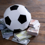 Bola de futebol apoiada em pilhas de notas de euro e dólar americano, ilustrando apostas esportivas, manipulação de resultados, riscos de integridade em jogos de azar e regulamentações de apostas esportivas. Pesquisa sobre manipulação de resultados na Noruega mostra lacunas nos relatos de manipulação esportiva em meio a preocupações globais de integridade