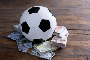 Bola de futebol apoiada em pilhas de notas de euro e dólar americano, ilustrando apostas esportivas, manipulação de resultados, riscos de integridade em jogos de azar e regulamentações de apostas esportivas. Pesquisa sobre manipulação de resultados na Noruega mostra lacunas nos relatos de manipulação esportiva em meio a preocupações globais de integridade