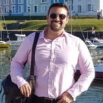 Andrew Fearon parado do lado de fora em frente a um porto, que contém vários barcos. Atrás do porto há um grande pedaço de grama e casas coloridas. Ele está usando óculos escuros, uma camisa rosa e tem uma bolsa do lado esquerdo. Ele está sorrindo e olhando para a câmera.