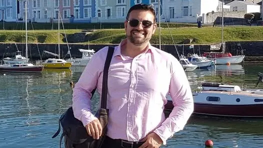 Andrew Fearon parado do lado de fora em frente a um porto, que contém vários barcos. Atrás do porto há um grande pedaço de grama e casas coloridas. Ele está usando óculos escuros, uma camisa rosa e tem uma bolsa do lado esquerdo. Ele está sorrindo e olhando para a câmera.