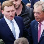 O líder do partido reformista do Reino Unido, Nigel Farage, reage depois que uma mulher jogou uma bebida nele. À sua esquerda está um segundo homem, vestido de terno com camisa branca e gravata azul.