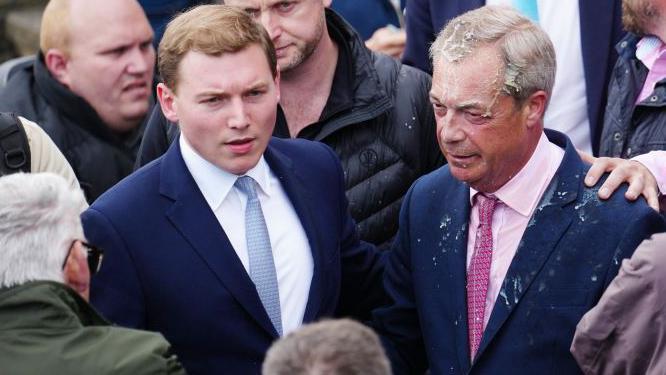 O líder do partido reformista do Reino Unido, Nigel Farage, reage depois que uma mulher jogou uma bebida nele. À sua esquerda está um segundo homem, vestido de terno com camisa branca e gravata azul.